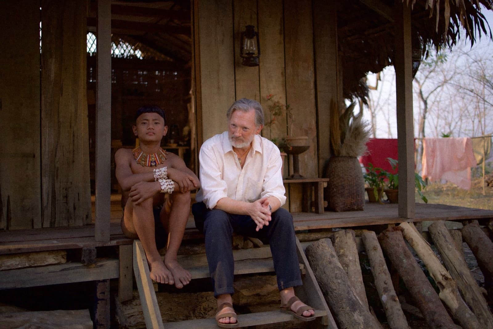 A Konyak boy in traditional necklace sits beside a grey-haired white missionary on the steps of a wooden house