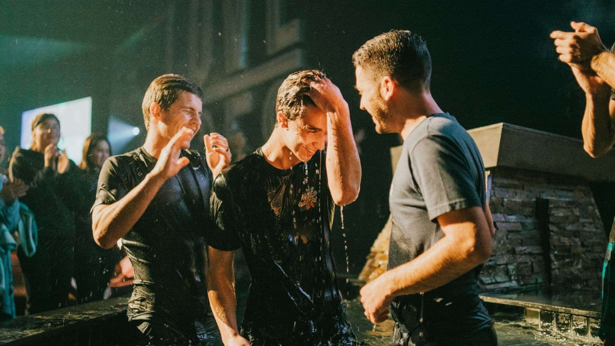 A young man stands soaked in a large stock tank of water, head bowed and water running off him, having just been baptised. Two men stand with him, one with a hand on his head, while people watch from behind.