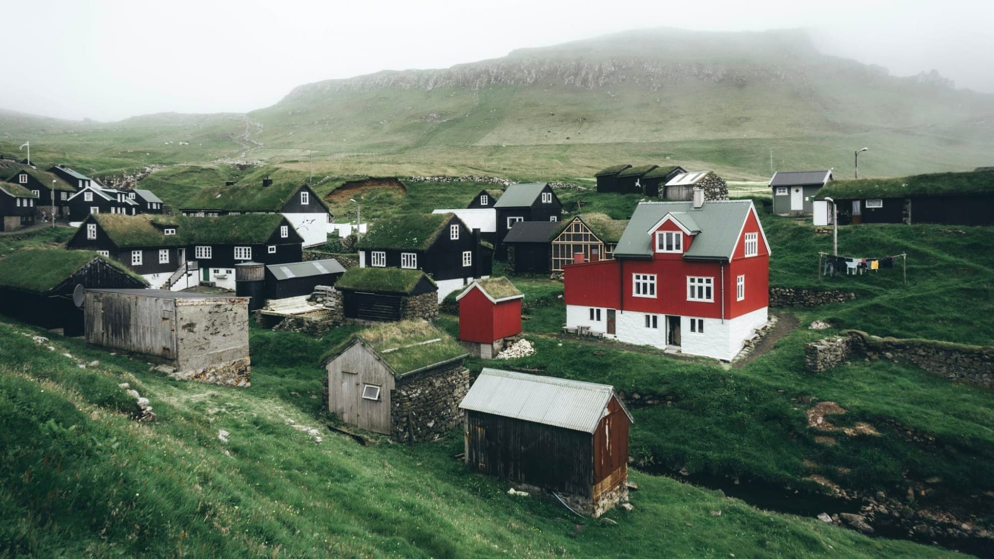 A Faroese village on a green hillside under low cloud: black timber houses with turf roofs, a red and white house among them, stone walls and a bare ridge behind.