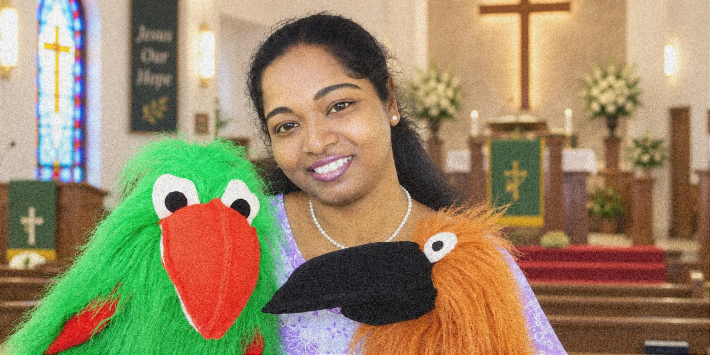 Mary Sujani holding two hand puppets, a green bird and an orange one, smiling at the camera