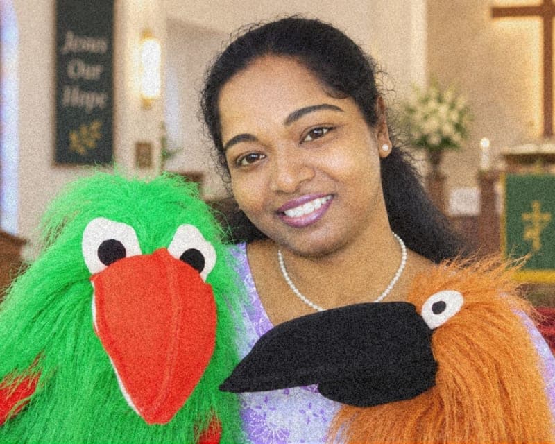 Mary Sujani holding two hand puppets, a green bird and an orange one, smiling at the camera