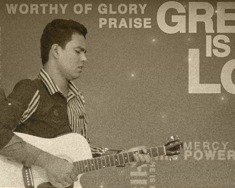 Ricky Biswasi playing a Korg keyboard during a service, with the congregation and a lit cross behind him