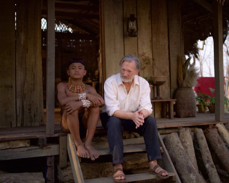 A Konyak boy in traditional necklace sits beside a grey-haired white missionary on the steps of a wooden house