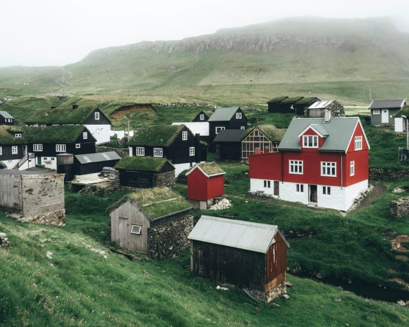A Faroese village on a green hillside under low cloud: black timber houses with turf roofs, a red and white house among them, stone walls and a bare ridge behind.